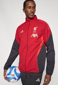 Red and black athletic jacket with a zip front, featuring the L.F.C. logo and AXA branding, paired with a blue and white soccer ball.