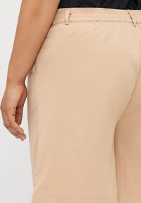 Pantalones beige de corte relajado con cintura ajustable y textura suave, con un bolsillo lateral visible cerca de la cadera y un corte recto.