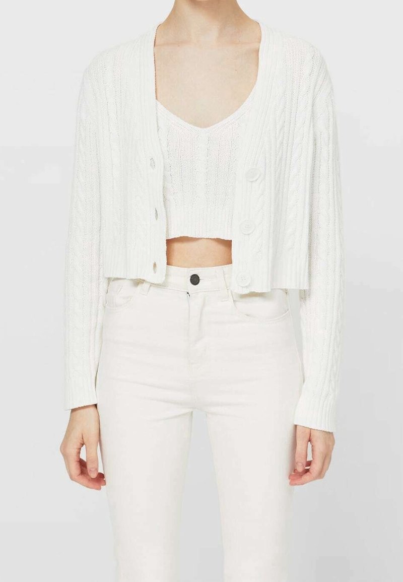 Bílý zkrácený kardigan se vzorem copánkového pletení, zdobený velkými knoflíky, nošený přes odpovídající pletený top a doplněný bílými džínami.