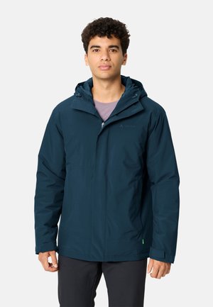 Giovane uomo che guarda verso davanti, indossa una giacca con cappuccio blu scuro e pantaloni neri, in piedi contro uno sfondo chiaro e uniforme.