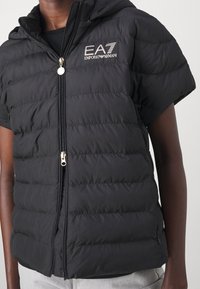 Černá péřová vesta s kapucí, má přední zip, krátké rukávy a logo EA7 Emporio Armani na levé straně hrudi. Hladká textura.