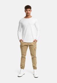 Jeune homme debout, les mains dans les poches, portant une chemise blanche à manches longues, un pantalon cargo beige et des baskets blanches sur un fond uni.