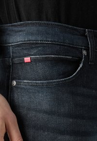 Jean en denim bleu foncé avec une petite étiquette rouge portant le logo « HUGO » sur la poche avant. Le tissu est lisse avec un léger éclat.