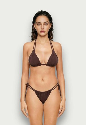 Mujer con cabello oscuro y rizado que lleva un bikini de hilo marrón con top halter y braguitas con lazos a los lados sobre un fondo liso y claro.