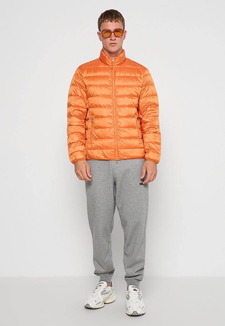 Veste rembourrée orange avec des coutures horizontales, col haut et fermeture éclair à l'avant. Portée avec un pantalon de survêtement gris. Modèle portant des lunettes de soleil et des baskets.