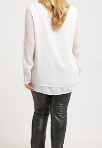 Blusa blanca de manga larga con una textura suave, mangas transparentes y un dobladillo redondeado, combinada con pantalones de piel negros.