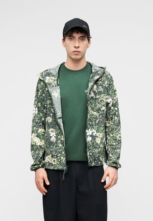 Young man wearing a black cap, green patterned hooded jacket over a green T-shirt, and black pants, standing against a plain light background.