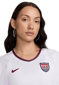 Maglia bianca con accenti rossi e blu, caratterizzata da un colletto rotondo e dal logo ricamato della US Soccer. Il materiale sembra liscio, probabilmente sintetico.