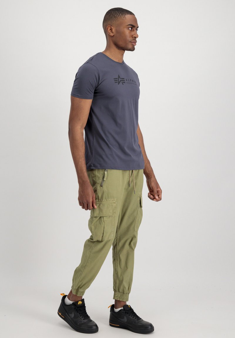 Szara koszulka z krótkim rękawem z czarnym logo na piersi, połączona z oliwkowymi spodniami cargo i czarnymi sneakersami.