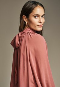 Indumento fluido di un delicato rosa, con cappuccio e dettaglio a fiocco, texture morbida e vestibilità ampia, che cade elegantemente sulla spalla.