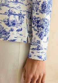 Langärmliges Oberteil mit einem blauen und weißen Landschaftsdruck, der Häuser und Palmen zeigt. Gerippte Textur mit seitlichen Schlitzen und sichtbarem Markenlabel.