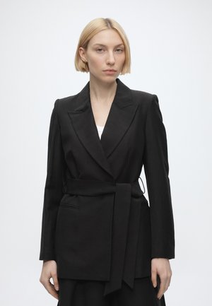 Femme aux cheveux blonds courts portant un blazer noir ceinturé sur un haut blanc devant un fond blanc uni.