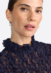 Blusa in pizzo navy con un colletto alto e arricciato, caratterizzata da un motivo floreale testurizzato e un tessuto trasparente, completata da piccoli orecchini a cerchio.