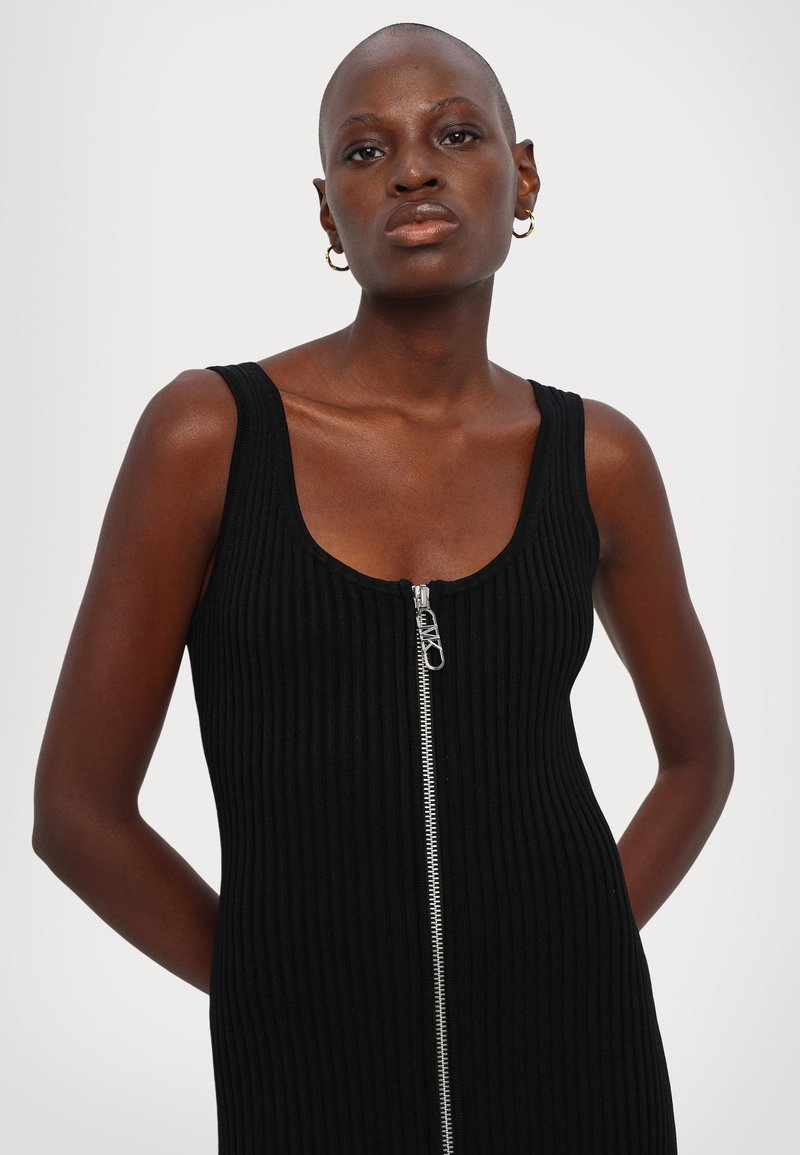Robe noire à côtes, avec un col en scoop et un détail de fermeture éclair à l'avant. Texture lisse, forme ajustée avec des bretelles larges, design longueur genou.