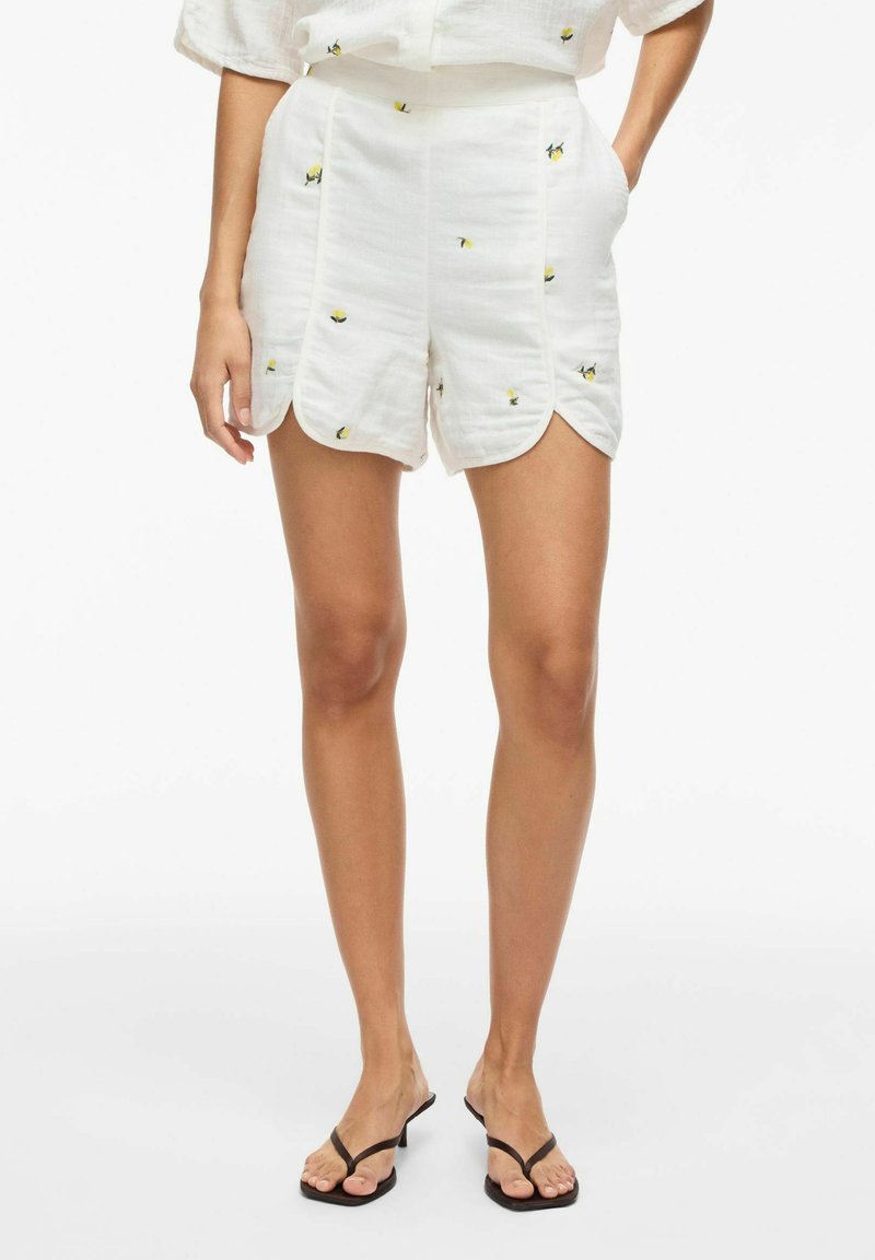 Short blanc taille haute avec des ourlets festonnés et un petit motif floral jaune, porté avec des sandales noires à lanières, mains dans les poches.