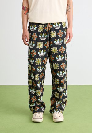 Nogi w luźnych czarnych spodniach z wielokolorowymi geometrycznymi i kwiatowymi wzorami, białe trampki i częściowo widoczna jasnobeżowa bluzka.