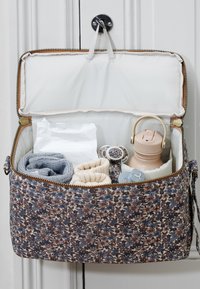 Baby taske med blomsterprint hængende åben med strikket tøj, babyvådservietter, to sutter og en lyserød træningskop indeni.