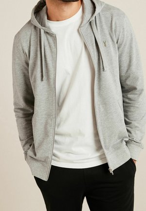 Lichtgrijze rits hoodie met een trekkoordcapuchon, met een voorzak, gedragen over een wit t-shirt en zwarte broek. Zachte textuur.