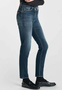 Jean en denim bleu foncé avec une coupe slim, présentant des accents dorés sur les boutons, une taille haute et un léger décoloration le long des jambes.