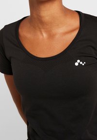 Svart atletisk t-shirt med rundhals, som har texturerade detaljer och en vit logotypdesign på vänster bröst. Mjuk, figurnära material.