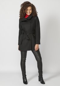 Cappotto nero su misura con cintura, colletto asimmetrico e tasche con zip. Indossato sopra leggings neri testurizzati e stivaletti con tacco.