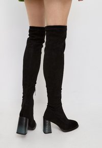 Bottes noires au-dessus du genou en daim, avec un talon bloc et un accent métallique strié, ainsi qu'une fermeture éclair sur le côté.