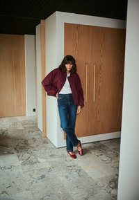 Veste bombardier bordeaux sur une chemise blanche, assortie à un jean droit bleu et des chaussures plates rouges. L'arrière-plan présente des meubles en bois.