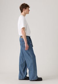 White short-sleeve t-shirt, paired with blue wide-leg jeans, featuring a brown label on the back pocket and a loose fit, worn with black shoes.