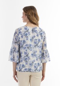 Blomstret bluse med knaplukning på ryggen og klokkeærmer. Stoffet er let og translucent, og viser blå og grå roser på en cremet baggrund.