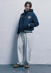 Námořnická varsity bunda s vyšívanými detaily, bílé pruhy na manžetách a lemu, v kombinaci se šedými tepláky a hnědými botami s modrými tkaničkami.