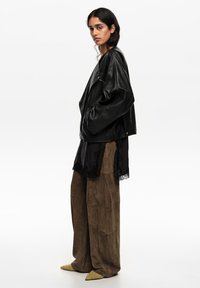 Naine seisab külgvaates, kandes üle suurusega musta nahktagi, musta pitsiga äärega topi, laiu pruune seemisnahkpükse ja teravate varvastega beeže kingi.