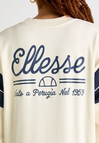 Krémová mikina s námorníckomodrým textom a akcentmi. Obsahuje logo "Ellesse", horizontálne pruhy a okrúhlu grafiku pod textom.