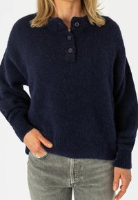 Maglione oversize lavorato a maglia blu navy con finitura testurizzata, caratterizzato da uno scollo tondo e tre bottoni viola. Abbinato a jeans grigi.