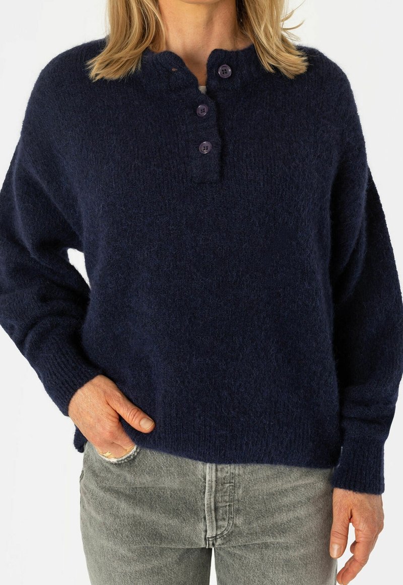 Maglione oversize lavorato a maglia blu navy con finitura testurizzata, caratterizzato da uno scollo tondo e tre bottoni viola. Abbinato a jeans grigi.