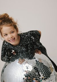 Schwarzes Paillettenoberteil mit gerafften Ärmeln, getragen von einem Kind, das auf einer reflektierenden Diskokugel sitzt. Glatte, glänzende Textur und kreisförmiges Design.