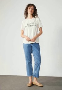 Λευκό βαμβακερό t-shirt με μαύρο κείμενο που λέει "LA VIE EST BELLE", συνδυασμένο με ανοιχτό μπλε τζιν και μπεζ φλατ παπούτσια σε ουδέτερο φόντο.