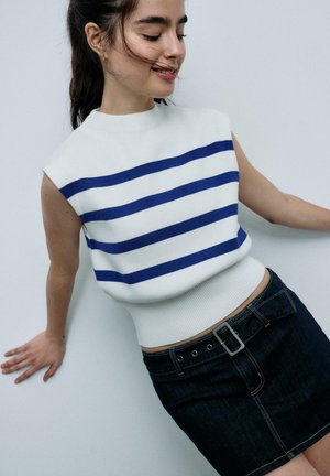 Frau lehnt sich an eine weiße Wand, trägt ein weißes ärmelloses Oberteil mit blauen Streifen und einen dunklen Jeansrock mit Gürtel.