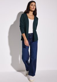 Tummanvihreä neuletakki valkoisen paidan päällä, yhdistettynä tummansinisistä leveälahkeisiin farkkuihin ja valkoisiin tennareihin, joissa on rento istuvuus ja teksturoitu kangas.