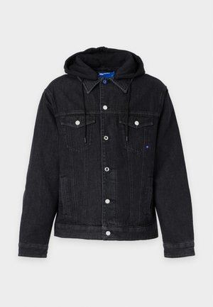 Jeune homme portant une veste en jean noire avec capuche, un t-shirt blanc, un jean noir et des baskets noires, debout avec une main dans la poche.