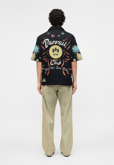 Άτομο που φοράει μαύρο κοντομάνικο μπλουζάκι με πολύχρωμα γραφικά και το κείμενο «Barrou Club Bad Vibes. Good Times» στην πλάτη, και μπεζ παντελόνι.