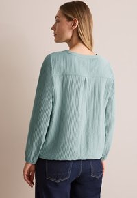Blusa lunga a maniche di colore verde menta, strutturata, con collo rotondo e piega sul retro, abbinata a jeans blu scuro con tasche visibili.