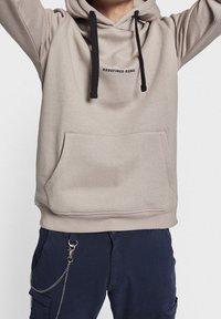 Beige pullover hoodie with a front pouch pocket, black drawstrings, and the text "REDEFINED REBEL" in black on the chest.