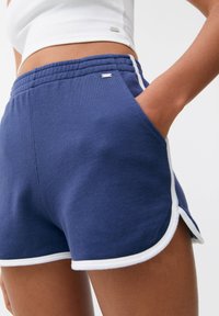 Shorts en coton bleu marine avec bordure blanche, taille élastique, poches latérales et un petit logo sur le devant. Texture douce, design sportif.