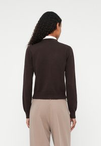 Pull en maille marron avec des manches longues, poignets et ourlet côtelés, il présente un col montant. Porté avec un pantalon beige clair, vu de dos.