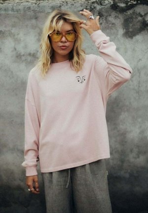 Młoda osoba o falowanych blond włosach, nosząca okulary z żółtymi szkłami, jasnoróżową bluzę z wzorem twarzy oraz szare spodnie, stoi na tle teksturowanej ściany.