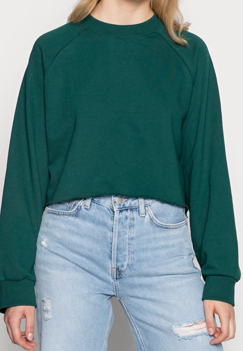 Femme portant un sweat-shirt court à manches longues vert foncé et un jean délavé taille haute bleu clair, debout, les bras détendus.