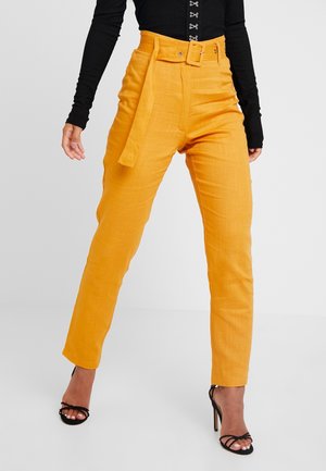 Frau trägt hoch taillierte, senfgelbe Gürtelhose, schwarzes langärmeliges Oberteil und schwarze, mit Riemchen versehene High-Heel-Sandalen, steht mit gespreizten Beinen.