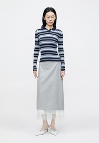 Žena s rovnými čiernymi vlasmi, oblečená v modrom pruhovanom tričku s dlhým rukávom, sivej midi sukni s bielou čipkovanou lemovkou a bielych špicatých podpätkoch.