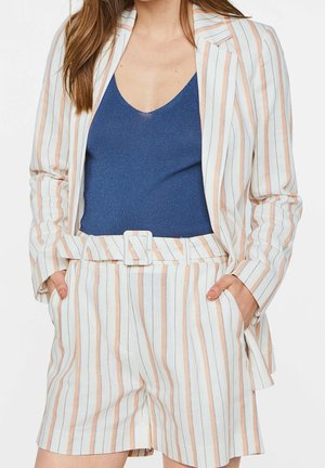 Žena v bílém blazeru a odpovídajících šortkách se sponou, které mají broskvově a černě pruhovaný design, a námořnickém modrém topu bez rukávů.