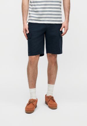 Férfi sötétkék cargo rövidnadrágban, fehér zokniban, barna bőr hajócipőben, valamint fehér pólóban, amelyen tengerészkék vízszintes csíkok találhatók.